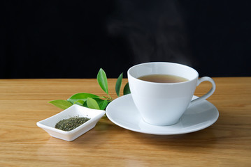 Green tea in a white cup put on a white saucer with hot steam over the cup. Seen on wooden table on black background. Green tea leaf ingredient in white plate and green leaf also put near the cup.