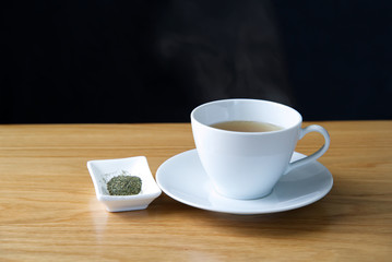 Green tea in a white cup put on a white saucer with hot steam over the cup. Seen on wooden table on black background. Green tea leaf ingredient in white plate also put near the cup.