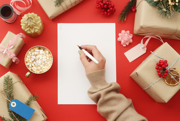 Female hand writes on a white sheet of paper among Christmas gifts, decorations next to a cup of cocoa with marshmallows on a red background. Top view, flat lay
