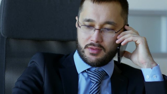 Medium shot of Asian man in glasses and business suit having phone conversation with colleague when working in office