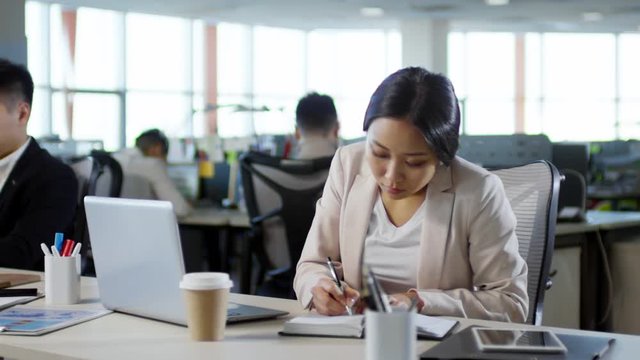 Tracking shot of concentrated young Asian woman taking notes in notebook and typing on laptop computer when sitting at desk in office, her colleagues working in the background