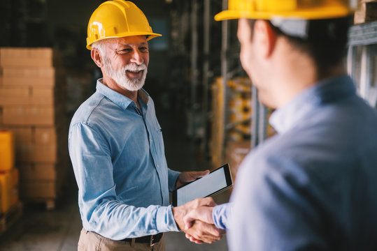 Two satisfied business man standing in warehouse with helmets on their head. Shaking hands and closing good job cooperation.