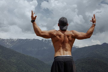 Rear view of muscular bodybuilder with naked tanned torso, arms up outstretched looking at...