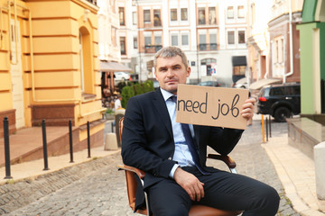 Businessman holding piece of cardboard with text NEED JOB while sitting in office armchair outdoors