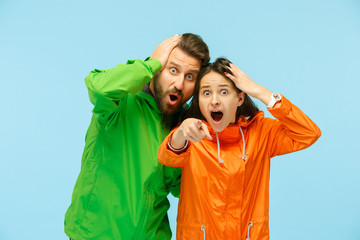The young surprised couple pointing to camera and posing at studio in autumn jackets isolated on blue. Human negative emotions. Concept of the cold weather. Female and male fashion concepts