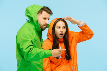 The young surprised couple pointing to left and posing at studio in autumn jackets isolated on blue. Human negative emotions. Concept of the cold weather. Female and male fashion concepts