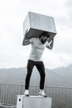 Monochrome Shot Of Crossfit Man Doing Squats With Weighting On Wooden Block, Holding Another Block In Hand, Outdoor Crossfit Work Out Over Mountain Nature Background.