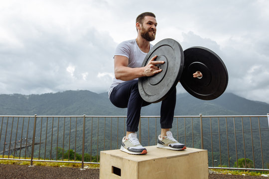Motion outdoor shot of cross-fit sportsman doing plyometric jumping squats with weights at outdoor functional workout over mountain and sky background.