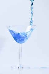 Cocktail with blue liquid in glass. Glass with blue water pouring with liquid with splashes and drops. Martini glass filling with alcohol with splashes on white background. Refreshing drink concept.
