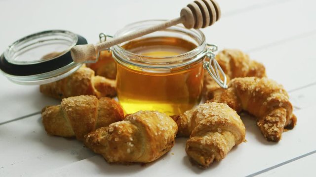 Glass jar full of honey with wooden spindle surrounded by fresh croissants on white wooden background 