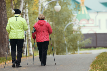 Two elderly women are doing Scandinavian walking in the park. Autumn. Back angle