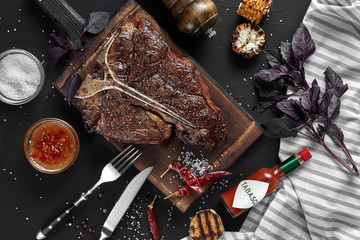Succulent grilled large t-bone steak garnished with herbs, corn and salt with fork and knife beside it on cutting board