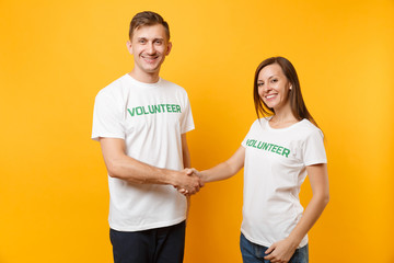 Portrait of young colleagues couple in white t-shirt with written inscription green title volunteer isolated on yellow background. Voluntary free work, assistance help, charity grace teamwork concept.