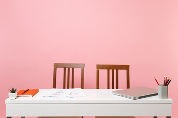 Office light interior of workplace, table with modern laptop pc computer documents orange notebook pencils flower and chairs on pastel pink background. Copy space advertising youth co working concept.