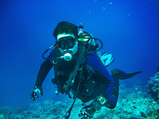 Scuba diver is underwater. He is wearing in full scuba-diving equipment: mask, regulator, BCD, fins. Diver is on the blue water background and corals are on the bottom.