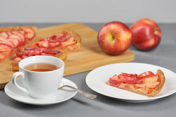 A piece of apple pie on a plate. Next to a cup of tea. In the background are apple pie and apples. Gray background. Close-up.