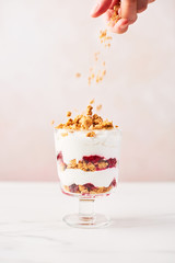 Hand sprinkling granola over healthy raspberry yogurt parfait in a glass on white marble table over pink background with copy space.