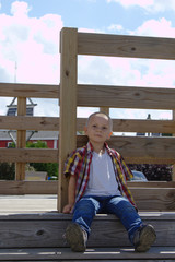 Cute Young School Age Boy With Short Blond Hair Brown Eyes and Red and Yellow Plaid Shirt Sitting on a Wooden Platform In Front of a Farm