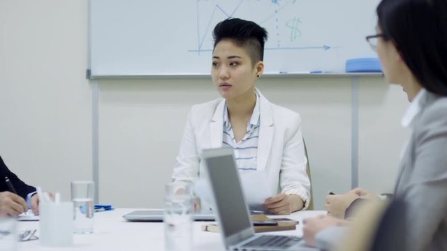 Medium shot of young Asian female boss sitting at desk in conference room and criticizing ineffective employees at meeting