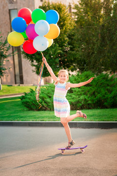 Happy little girl skating on a scateboard with large bunch of helium filled colorful balloons
