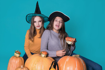 Fototapeta premium Portrait of two happy young women in black witch halloween costumes on party over blue background. The concept of Halloween, funny faces. Teenage girls with pumpkin and attributes of the holiday.