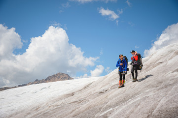 Fototapeta premium Two tourists, a man and a woman with backpacks and cats on their feet, stand on the ice in the background of the mountains of the sky and clouds. communication in the mountains