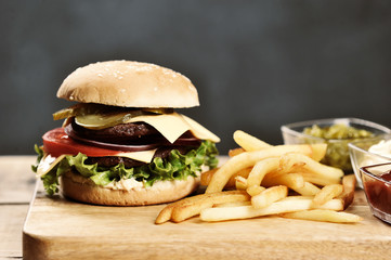 Double cheeseburger and french fries on a light wooden table. Burger filling consists of two steaks, vegetables, cheese and sauce.  Dark background. Close-up.