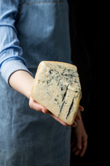 Woman in apron holding piece of blue cheese Gorgonzola on dark background