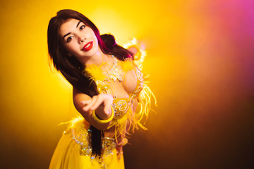 Tempting sexy traditional oriental belly dancer girl dancing on yellow neon smoke background. Woman in exotic costume with feathers sexually moves her semi-nude body