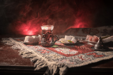 Arabian tea in glass with eastern snacks on a carpet on dark background with lights and smoke. Eastern tea concept. Empty space.
