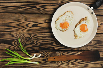 Two fried eggs in metal beige pan near dirty spatula and fresh green onion on old wooden brown table. Top view with copy space