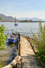 Naklejka premium Barca blu ancorata al molo di un lago con isola e montagne sullo sfondo, Isola dei Pescatori, Lago Maggiore, Italia