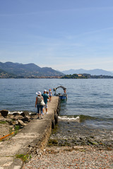 Veduta di un lago con turisti su un molo diretti verso una barca, isola e montagne sullo sfondo, Lago Maggiore, Isola dei Pescatori, Piemonte, Italia