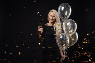 Obraz na płótnie Canvas Brightfull expressions of happy emotions of amazing blonde girl celebrating party on black background. Luxury black dresses, smiling, a glass of champagne, golden tinsels, balloons, long curly hair