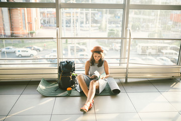 Waiting, delayed transport in the terminal of the airport or train station. Young caucasian woman in dress and hat sits on tourist rug with backpack near window to room and looks into tourist map