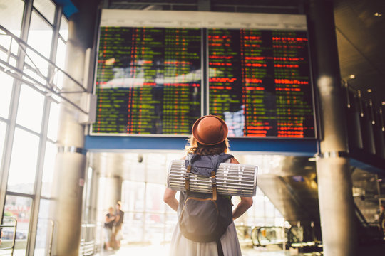 Theme travel public transport. young woman standing with back in dress and hat behind backpack and camping equipment for sleeping, insulating mat looks schedule on scoreboard airport station