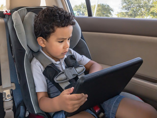 A little boy holds a tablet while strapped in his car seat.