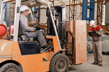 the storekeeper marks the arrival on the tablet. man driving a forklift through a warehouse in a factory. driver in uniform and protective helmet. the concept of logistics and storage