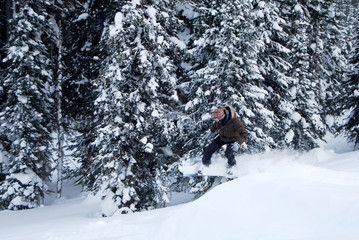 Male snowboarder on the snowy slope on sunny winter day. Young sportsman jumping, spraying snowflakes and flying on background of trees covered with snow.