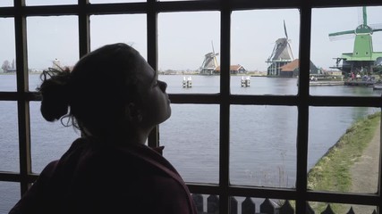 Young woman opposite window looking at typical holland landscape dutch village on the rewerbank with old-fashioned windmills