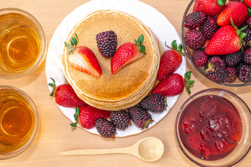Tasty breakfast. Pancakes with fruit, strawberries and blackberries (mora), poured syrup (honey) on a wooden tray. Two cups of tea.