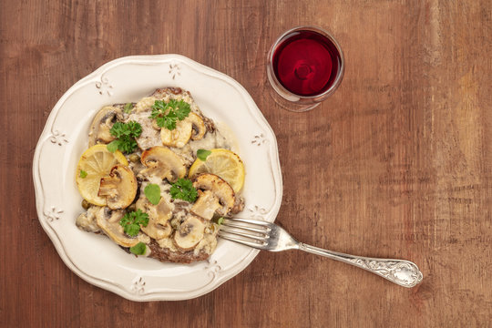 Overhead photo of scallopine di vitello, veal scallopini, with a mushroom sauce, lemons, and parsley, on a dark rustic texture with red wine and copy space