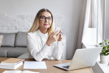 Age, business, job and technology concept. Portrait of successful modern elderly woman psychologist wearing white shirt and eyeglasses typing sms using cell phone, sitting at her workplace