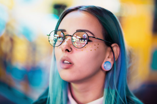 Millennial pretty girl with unusual dyed hairstyle smoking cigarette in amusement park at night. Blue hair, golden sequins as freckles,nose piercing. Mysterious hipster.