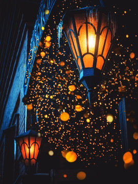 A Magical Old Street Lanterns Shines on the Street at Night. Many bright lights around. Vintage Old Street Classic Iron Lanterns On The House Wall. Christmas or Halloween Magic Fairy Lanterns