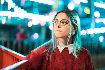 Hipster girl with blue dyed hair and sequins as freckles. Woman in red clothing and nose piercing, transparent glasses, ears tunnels, unusual hairstyle in amusement park - obrazy, fototapety, plakaty