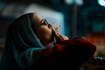 Millennial cool pretty girl with unusual dyed hairstyle smoking cigarette near glowing neon wall at...