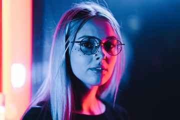 Millennial enigmatic pretty girl with unusual dyed hairstyle near glowing neon wall at night. Blue hair, golden sequins as freckles,nose piercing. Mysterious hipster teenager in glasses