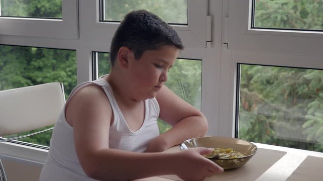 Bored little fat boy dining in the kitchen, eating a tablespoon of soup, concept childhood obesity and binge eating