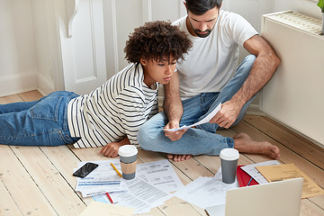 Mixed race graphic designers work remotely at home. Male and female students hold papers and check data on laptop computer, create coworking space on floor in modern apartment, drink beverage